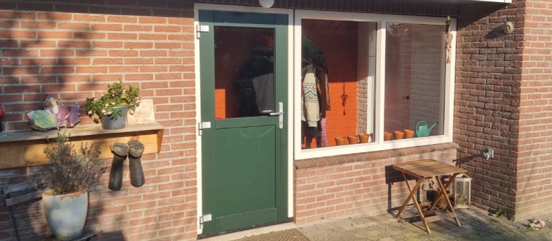 Wil je gemak? Kies dan voor kunststof kozijnen. Rood bakstenen muur met een groene deur en witte kozijnen.