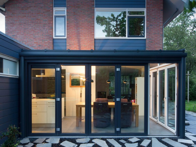 Een buitenfoto van de aanbouw van een modern huis met grote, glazen vouwdeuren en een stenen pad in de achtertuin.