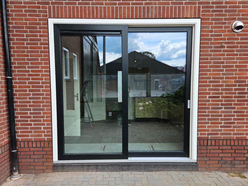 Een kunststof schuifpui met een zwart frame en een wit buitenkozijn, geplaatst in een rode bakstenen muur. De pui weerspiegelt de lucht en de overkant van de tuin.